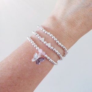 May include: Three silver beaded bracelets with a butterfly charm. The bracelets are stacked on a wrist and feature small silver heart charms.