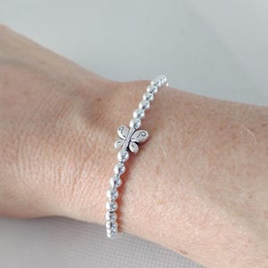 May include: A silver beaded bracelet with a butterfly charm. The bracelet features small, round beads strung together, with a detailed butterfly charm at the center. The butterfly has intricate wing designs. The bracelet is worn on a wrist.