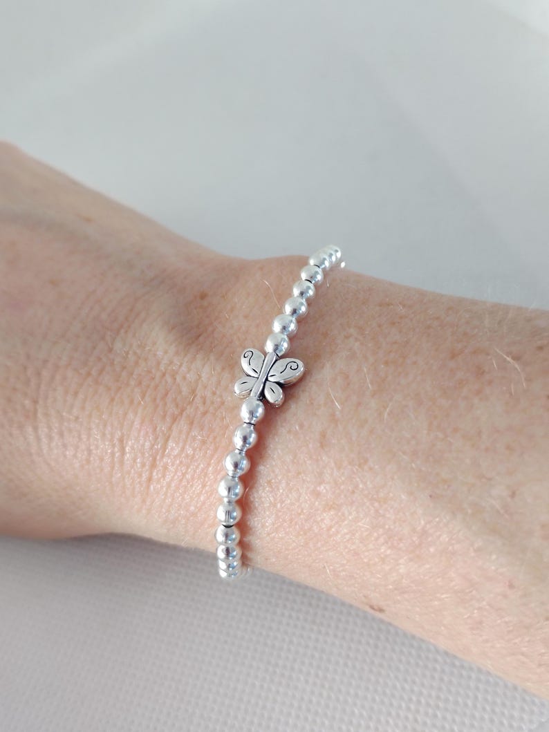 May include: A silver beaded bracelet with a butterfly charm. The bracelet features small, round beads strung together, with a detailed butterfly charm at the center. The butterfly has intricate wing designs. The bracelet is worn on a wrist.