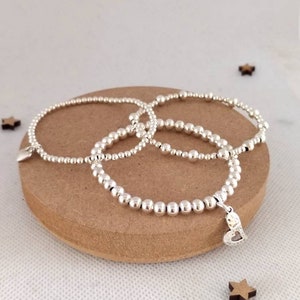 May include: Three silver beaded bracelets with a heart charm on one of the bracelets. The bracelets are stacked on a brown surface.