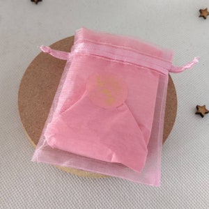May include: A pink organza gift bag with a gold sticker that says "Little Things" on it. The bag is tied with a pink ribbon.