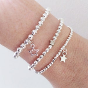 May include: Three silver bead bracelets with star charms. The bracelets are stacked on a person's wrist.