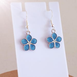 May include: A pair of silver earrings with a flower design. Each flower has five turquoise enamel petals. The earrings are displayed on a white stand. The earrings have a hook closure. The earrings are suitable for pierced ears.