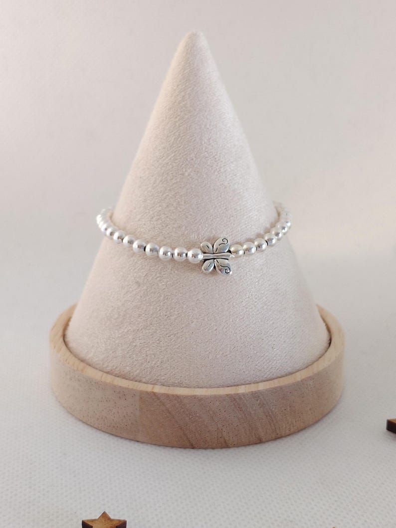 May include: A silver beaded bracelet with a butterfly charm. The bracelet is displayed on a light beige cone-shaped stand with a wooden base. The butterfly charm is centered on the bracelet.