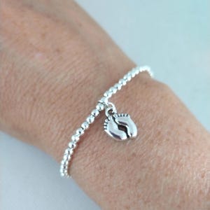 May include: A silver bracelet featuring small, round beads and a charm depicting baby footprints. The charm is suspended from the bracelet and has a detailed design. The bracelet is worn on a wrist.