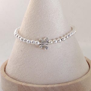 May include: A silver beaded bracelet with a butterfly charm. The bracelet is displayed on a cream-colored cone-shaped stand with a wooden base. The butterfly charm is centered on the bracelet.