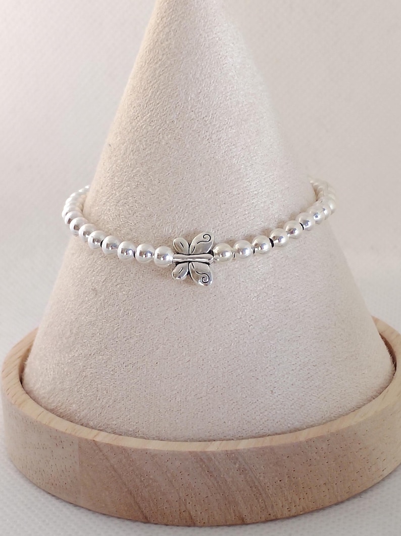 May include: A silver beaded bracelet with a butterfly charm. The bracelet is displayed on a cream-colored cone-shaped stand with a wooden base. The butterfly charm is centered on the bracelet.