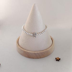 May include: A silver beaded ring with a four-leaf clover charm is displayed on a beige cone-shaped stand. The ring is made of small, round silver beads. The stand sits on a wooden base, with small wooden star decorations scattered around.