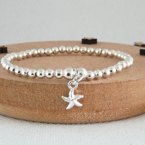 May include: A silver beaded bracelet with a small silver starfish charm. The bracelet is on a wooden surface.