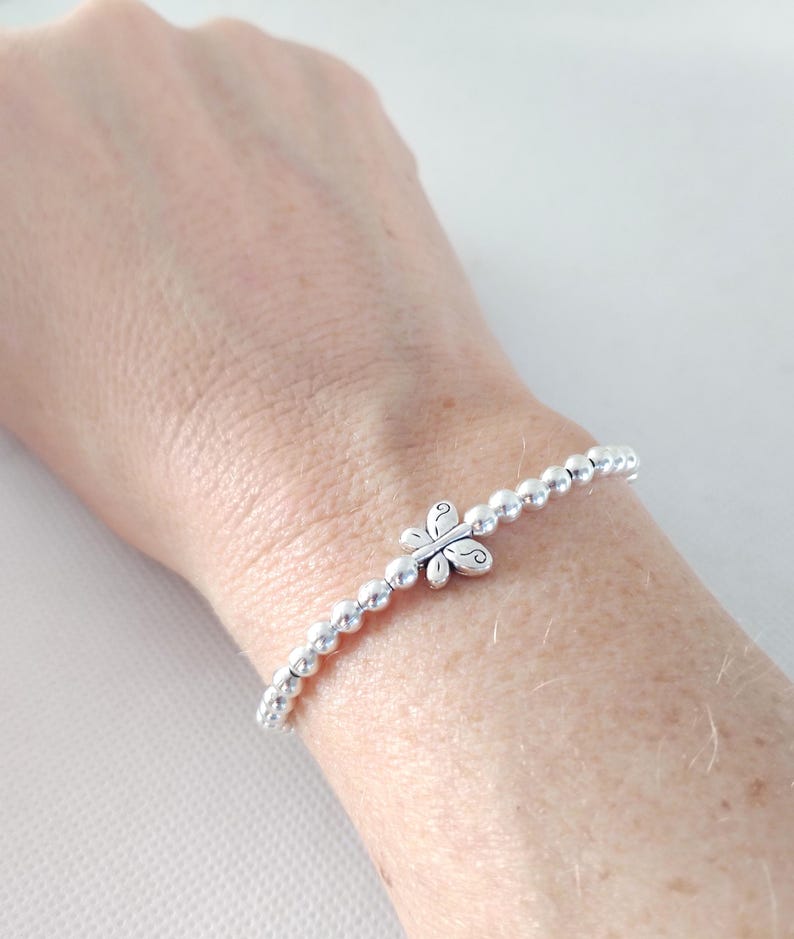 May include: A silver beaded bracelet with a butterfly charm. The bracelet features small, round beads strung together with an elastic band. The butterfly charm is centered on the bracelet and has a detailed design.