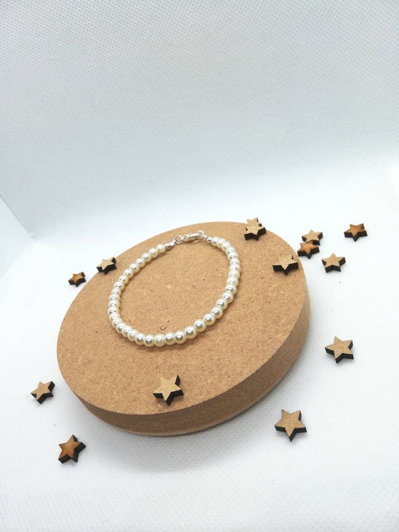 May include: A simple pearl bracelet with a silver clasp, displayed on a brown circular surface surrounded by small wooden stars.