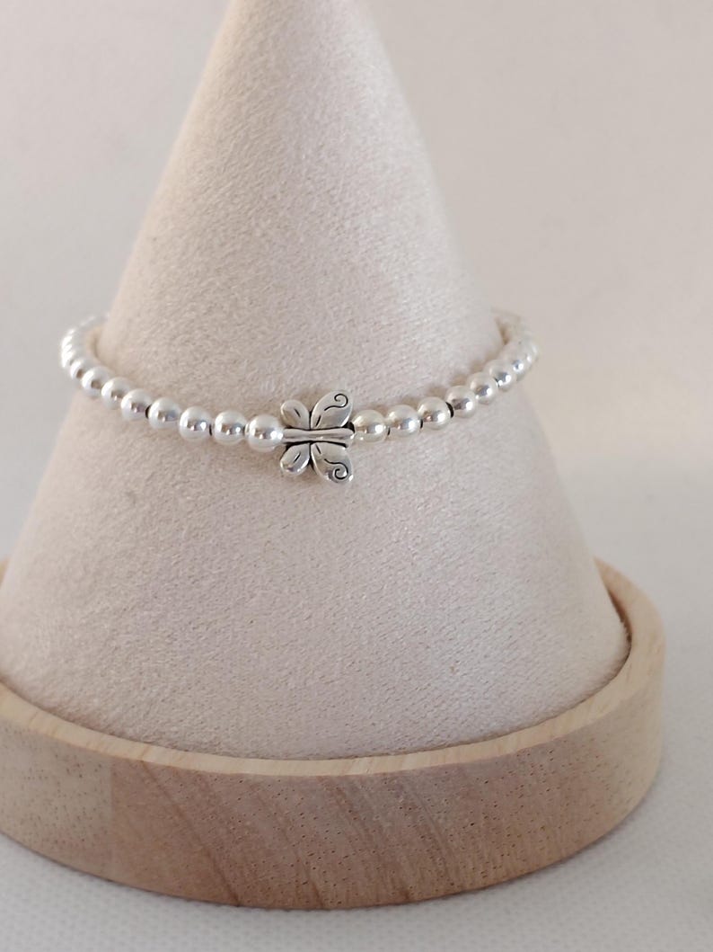 May include: A silver beaded bracelet with a butterfly charm. The bracelet features small, round silver beads and a detailed butterfly pendant at the center. The bracelet is displayed on a light-colored, cone-shaped stand.