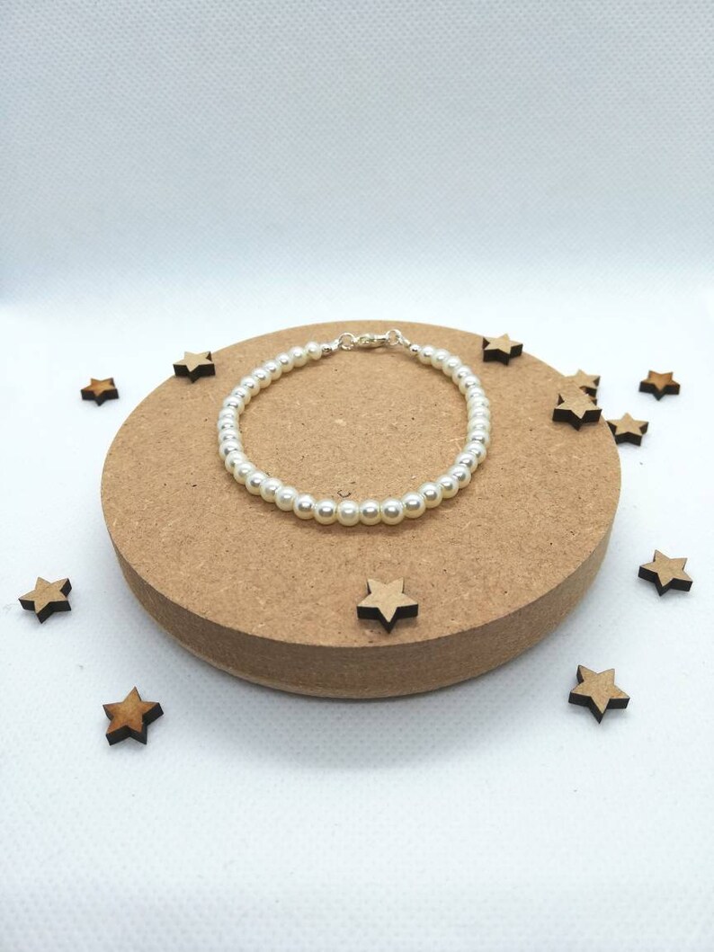 May include: A white pearl bracelet with a silver clasp. The bracelet is on a wooden surface with small wooden stars scattered around it.