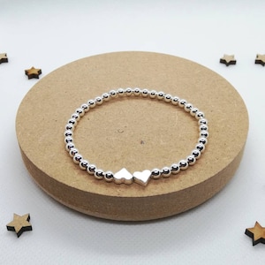 May include: A silver beaded bracelet with two silver heart charms. The bracelet is on a brown wooden surface.