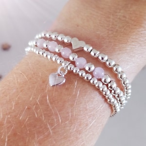 May include: Three silver beaded bracelets with pink beads and silver heart charms. The bracelets are stacked on a person's wrist.