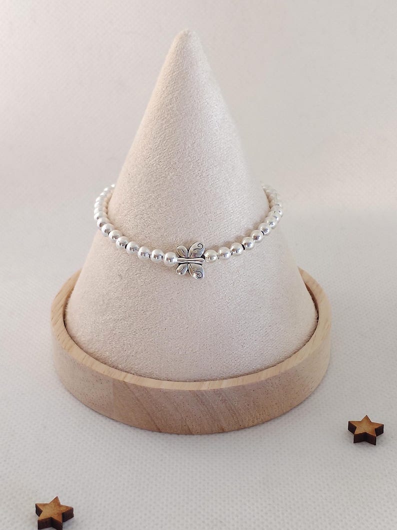 May include: A silver beaded bracelet with a butterfly charm. The bracelet is displayed on a white cone-shaped stand with a wooden base. Two small wooden star-shaped decorations are placed near the base.