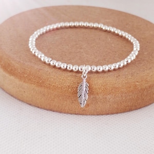 May include: A silver beaded bracelet with a small silver feather charm. The bracelet is on a wooden surface.