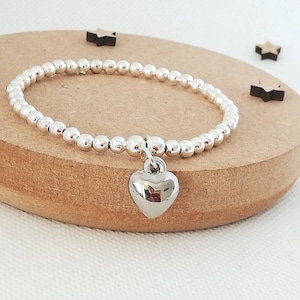 May include: A silver beaded bracelet with a silver heart charm. The bracelet is on a wooden surface.