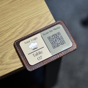 Puede incluir: Un porta-números de mesa de cuero marrón con un logotipo de gorro de cocinero blanco, el texto "Your Logo", "Table 07" y un código QR con el texto "Scan for Menu".