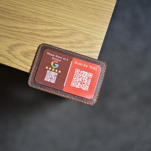 Puede incluir: Un porta tarjetas de cuero marrón con dos códigos QR. El código QR de la izquierda es para las reseñas de Google y dice "Por favor, déjanos una reseña". El código QR de la derecha es para WiFi y dice "Escanea para WiFi".