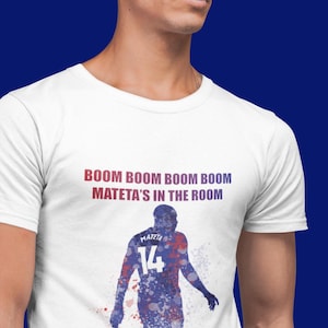 May include: White t-shirt with a blue and red watercolor silhouette of a soccer player with the number 14 on the back. The text "BOOM BOOM BOOM BOOM MATETA'S IN THE ROOM" is printed on the shirt.