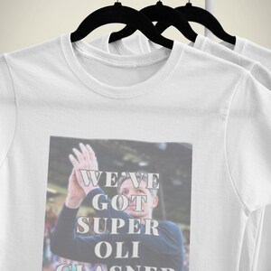 May include: White t-shirt with a graphic that says "WE'VE GOT SUPER OLI GLASNER". The image shows a man with his hands raised. The shirt is hanging on a black hanger.