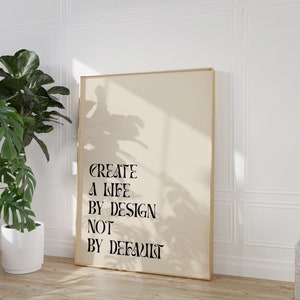 May include: A beige poster with a gold frame, hanging on a white wall. The poster features black text that reads "CREATE A LIFE BY DESIGN NOT BY DEFAULT".