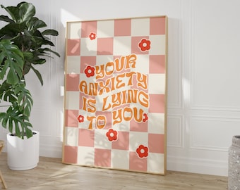 May include: A framed print with a retro design. The artwork features a pink and cream checkerboard pattern with the text "YOUR ANXIETY IS LYING TO YOU" in orange. Red flower illustrations are scattered across the print.
