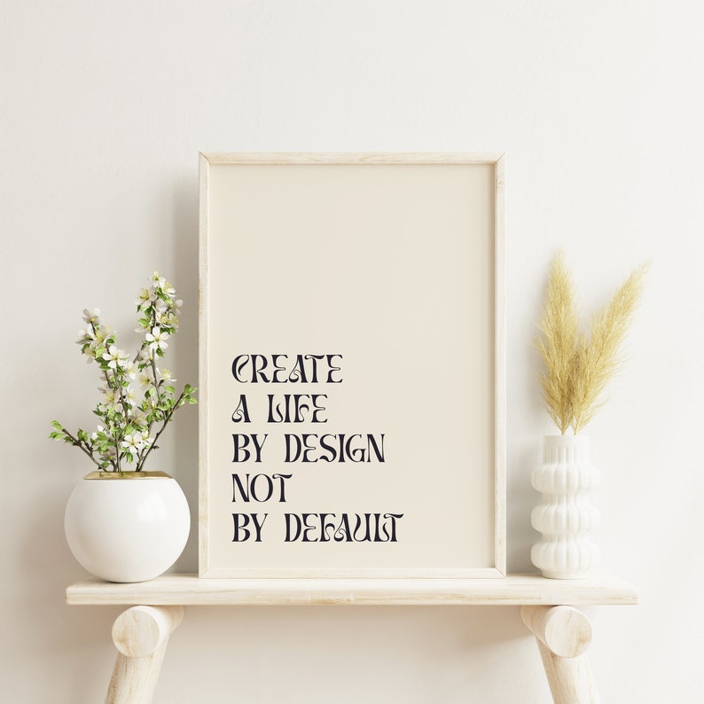 May include: A white framed print with the text "CREATE A LIFE BY DESIGN NOT BY DEFAULT" on a beige background. The print is on a white shelf with a white vase with dried grass and a white vase with a small flowering plant.