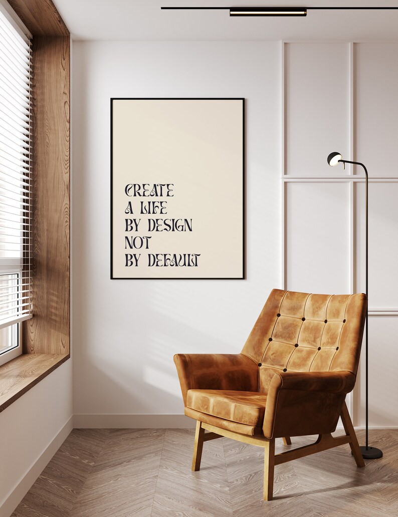 May include: A black framed poster with the text "CREATE A LIFE BY DESIGN NOT BY DEFAULT" hanging on a white wall in a room with a brown leather armchair and a floor lamp.