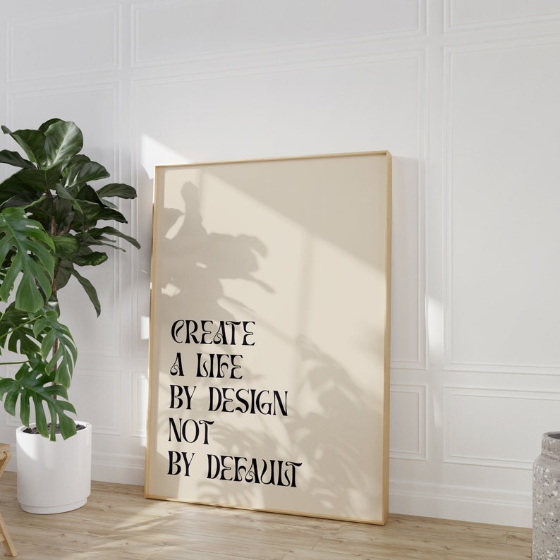 May include: A beige poster with a gold frame, hanging on a white wall. The poster features black text that reads "CREATE A LIFE BY DESIGN NOT BY DEFAULT".