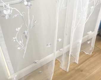 Könnte beinhalten: Transparenter weißer Vorhang mit gesticktem Blumen- und Blattmuster. Der Vorhang hat detaillierte Stickereien von Zweigen, Blumen und Blättern. Er hängt vor einem weißen Fensterrahmen und einem Holzboden.