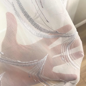 May include: Sheer white curtain fabric with embroidered gray feather-like designs. The delicate material is semi-transparent, allowing light to pass through. The embroidery adds texture and visual interest to the fabric, suitable for home decor.