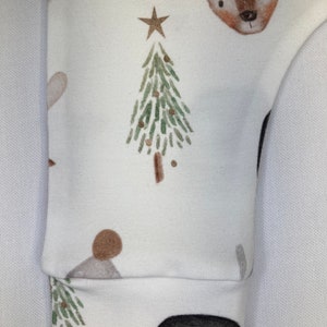 Puede incluir: Tela blanca con un patr&oacute;n repetido de un &aacute;rbol de Navidad, un ciervo, un ping&uuml;ino y una estrella. El pu&ntilde;o de la manga es visible, con la cara de un ping&uuml;ino en la parte inferior. El dise&ntilde;o presenta acentos marrones, verdes y dorados, creando un tema festivo.