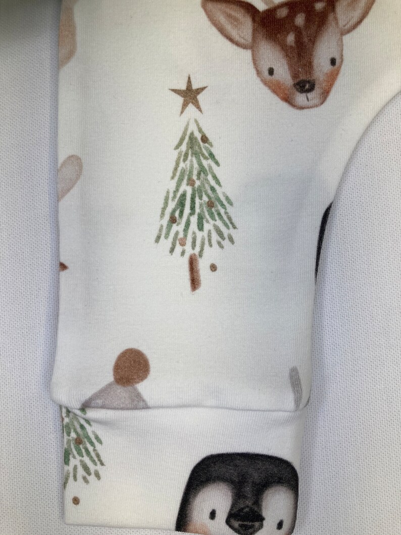 Puede incluir: Tela blanca con un patr&oacute;n repetido de un &aacute;rbol de Navidad, un ciervo, un ping&uuml;ino y una estrella. El pu&ntilde;o de la manga es visible, con la cara de un ping&uuml;ino en la parte inferior. El dise&ntilde;o presenta acentos marrones, verdes y dorados, creando un tema festivo.