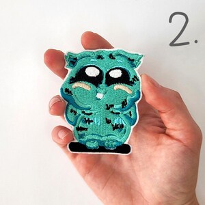 Puede incluir: Parche verde bordado para planchar de un personaje de dibujos animados con ojos grandes y una amplia sonrisa. El personaje lleva una camisa a rayas azules y blancas y tiene un contorno negro.