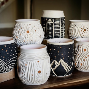 May include: A collection of handcrafted ceramic candle holders. The holders feature various designs, including floral patterns, cityscapes, and mountain scenes. Each holder has a unique texture and color scheme, with a mix of black and white, and a warm glow when lit.