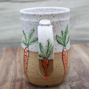May include: Ceramic mug with a speckled white top and tan base. The mug features hand-painted orange carrots with green tops. The handle also has a carrot design. A charming and unique piece for kitchen decor.