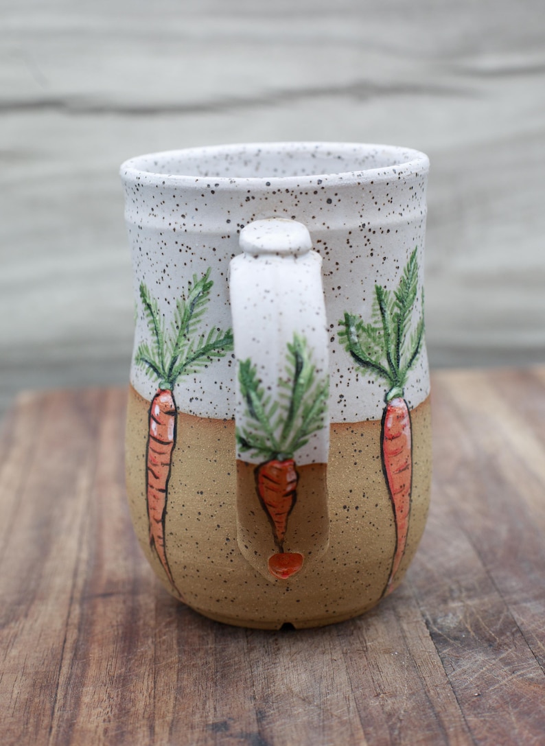 May include: Ceramic mug with a speckled white top and tan base. The mug features hand-painted orange carrots with green tops. The handle also has a carrot design. A charming and unique piece for kitchen decor.