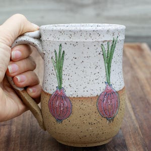 May include: A ceramic mug with a speckled white and brown finish. The mug features hand-painted red onion illustrations with green tops. The mug is held by a hand, showcasing its handle and design.