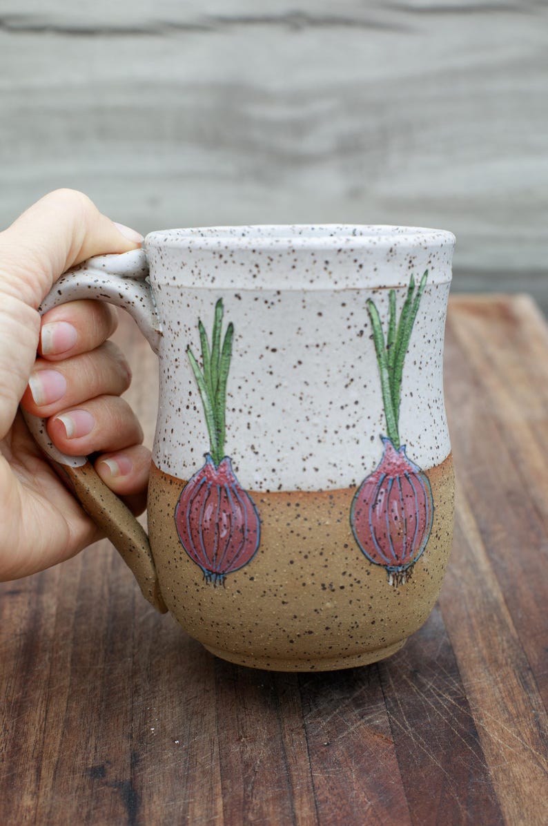 May include: A ceramic mug with a speckled white and brown finish. The mug features hand-painted red onion illustrations with green tops. The mug is held by a hand, showcasing its handle and design.