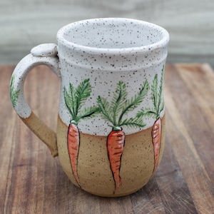 May include: A ceramic mug with a white speckled top and a tan base. The mug features hand-painted orange carrots with green tops. The handle is tan and white, and the mug is on a wooden surface.