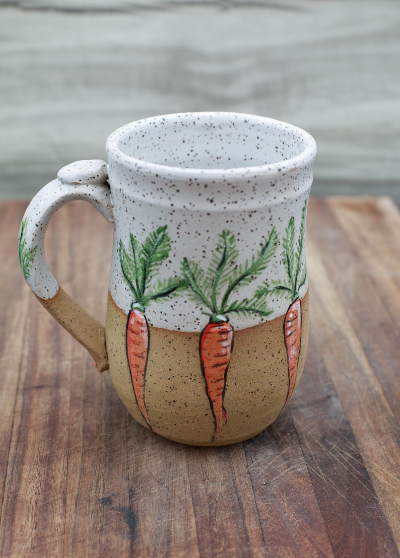 May include: A ceramic mug with a white speckled top and a tan base. The mug features hand-painted orange carrots with green tops. The handle is tan and white, and the mug is on a wooden surface.