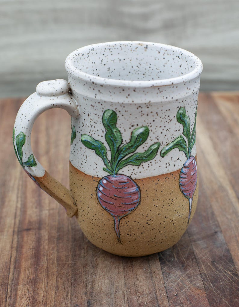 May include: Handmade ceramic mug with a white speckled top and a tan base. The mug features a hand-painted design of a red beet with green leaves. The handle is white with green accents. A unique and rustic kitchenware item.