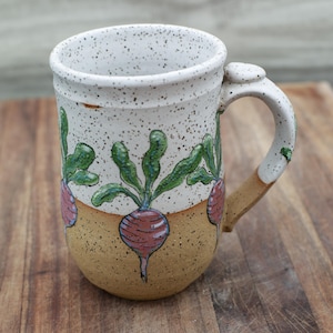 May include: Handmade ceramic mug with a speckled white exterior and a tan base. The mug features a hand-painted design of red radishes with green leaves. The handle is white with a tan accent.