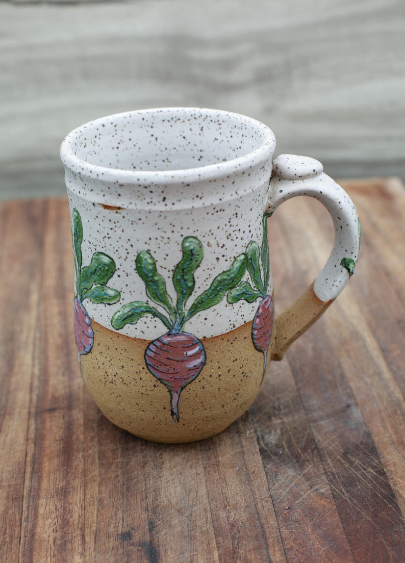 May include: Handmade ceramic mug with a speckled white exterior and a tan base. The mug features a hand-painted design of red radishes with green leaves. The handle is white with a tan accent.