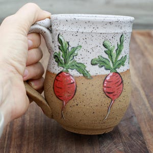 May include: A ceramic mug with a speckled white top and a brown base. The mug features hand-painted red radishes with green leaves. The handle is brown. The mug is held by a person.