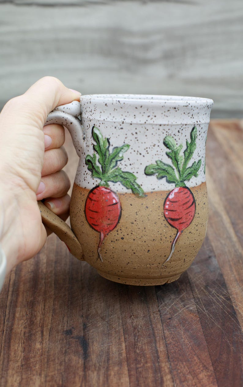 May include: A ceramic mug with a speckled white top and a brown base. The mug features hand-painted red radishes with green leaves. The handle is brown. The mug is held by a person.
