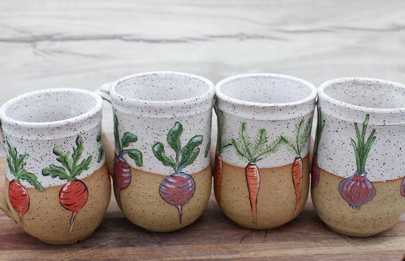 May include: Four handmade ceramic mugs with a speckled white top and tan base. Each mug features a different hand-painted vegetable: radish, carrot, and onion. The mugs have a rustic, farmhouse style.