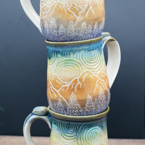 May include: Three ceramic mugs stacked vertically, each featuring a hand-carved relief design of mountains, pine trees, and swirling clouds. The mugs have a gradient glaze transitioning from blue at the rim to yellow and orange in the center.
